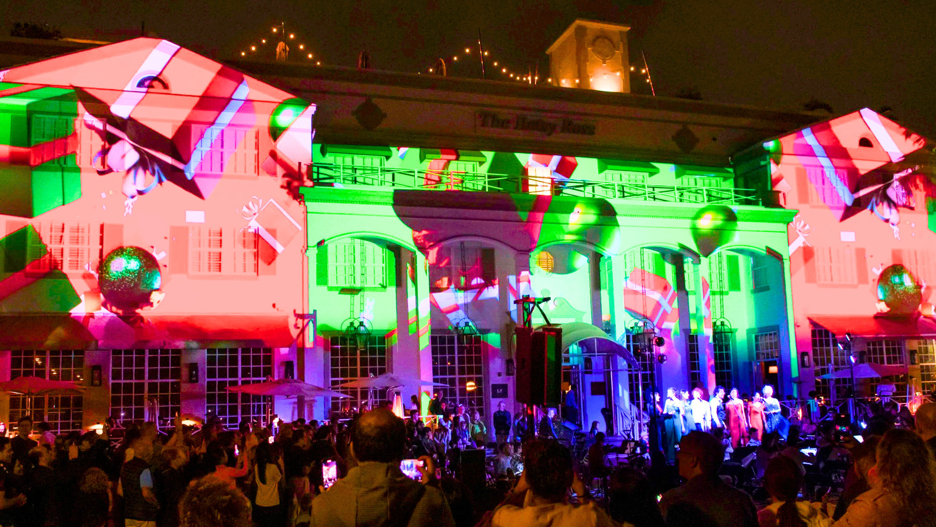 Photo of the Betsy Ross Hotel at night with Christmas designs projected onto the building exterior, a crowd of people watching and a classical music orchestra with singers performing in front of the hotel.