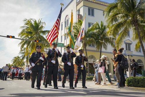 Miami Beach Fire Department members holding a golden axe with the Miami Beach Fire Department logo on it, an American flag, a Miami Beach flag and the Miami Beach Fire Department flag